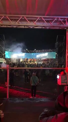 The video shows an outdoor nighttime concert with a large crowd gathered in front of a stage. A banner above the stage displays the word "KITONE" in large letters, with lights and smoke effects creating a festive atmosphere. On the stage, a group of people are visible, some of whom appear to be performers or DJs. The crowd is diverse, with individuals of various ages and genders. Many people are raising their hands in the air, dancing, and enjoying the music. The ground in front of the stage appears to be dirt or gravel, with some litter scattered around. The overall scene depicts a lively and energetic music event.
