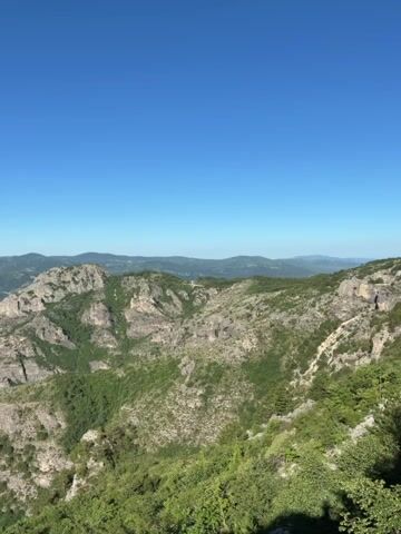 The video shows a panoramic view of a mountainous landscape under a clear blue sky. The foreground is dominated by lush green vegetation, including trees and shrubs, on a steep slope. Beyond this, the landscape unfolds into a series of rugged, rocky mountains covered in patches of greenery. The mountains appear weathered and show signs of erosion. In the distance, more rolling hills and mountains are visible, fading into a hazy horizon. The lighting suggests it is daytime, likely morning or afternoon, with bright sunlight casting shadows. There are no people or man-made objects visible in the frame. The scene is natural and undisturbed.