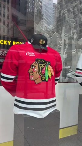 The video shows a store window display featuring Chicago Blackhawks hockey jerseys and caps on mannequins. The display includes a red jersey with a black and white striped trim and the Blackhawks logo on the back, and a white jersey with similar stripes and logo. A black cap with the Blackhawks logo is placed on top of the red jersey. In the background, a black and white mural depicts hockey players and fans. The store window reflects the street outside, showing buildings and people walking by. One shot shows a black jersey with the Blackhawks logo and a white cap. Another shot shows a person in a black shirt walking past the display. The video appears to be shot from a slight angle, looking into the store window.
