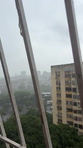 The video shows a view through a metal grate, looking out at a city skyline during what appears to be a rain shower. In the foreground, the bars of the grate are prominent and slightly out of focus. Beyond the grate, a tall, light-yellow apartment building dominates the frame. The building has many windows and air conditioning units visible. To the left and in the middle ground, lush green trees are seen, some of which are being affected by the rain. Further in the background, other buildings and a hazy cityscape are discernible, indicating a dense urban environment. The sky is overcast and gray, consistent with rainy weather. Small white specks, likely raindrops, are visible throughout the scene, especially against the sky and the building. The overall impression is one of a rainy day in a city, viewed from an elevated position.
