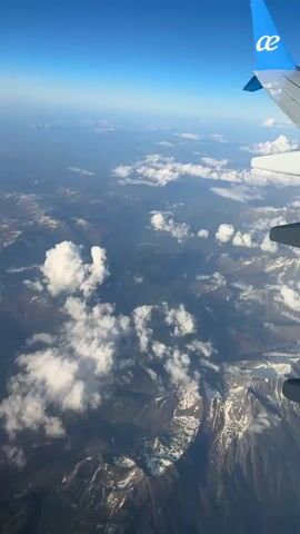 The view from an airplane window shows snow-covered mountains and valleys partially obscured by clouds under a bright blue sky. The wing of the airplane is visible, marked with a blue logo. The landscape below is a mix of dark mountain ranges and lighter patches of snow and clouds. The horizon is slightly hazy.