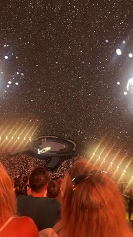 The video shows a concert audience facing a stage. Above the stage, a large projection displays a performer wearing a white hoodie and a white baseball cap, superimposed against a background of stars. The performer is gesturing with their right hand. In the foreground, the backs of audience members' heads are visible, with one person's ginger-colored hair prominent. Stage lighting is visible on either side of the stage. The overall impression is of a large-scale event with a projected visual element.