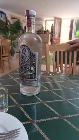 The video opens with a close-up shot of a bottle of vodka on a table. The bottle has a label with an image of a man in a suit and Cyrillic text. There are two glasses on the table, one larger and one smaller with a gold rim. A white plate with silverware is in the foreground. The background shows a restaurant interior with wooden chairs and tables, decorated with tinsel and paper banners. The walls are made of white brick. Sunlight streams in from the right side of the frame. The camera pans slightly to the left, revealing more of the restaurant and a person's hand resting on a chair. Another shot shows a pack of cigarettes on the table. The scene appears to be a relaxed setting, possibly a restaurant or a private dining area.