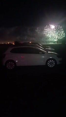 The video captures a nighttime scene featuring a white Volkswagen Polo parked in the foreground, with another vehicle behind it. The background is dominated by a fireworks display, with several bursts of white and gold explosions illuminating the dark sky. The fireworks appear to be set off at a distance, possibly in an open field or park. The overall atmosphere is festive, suggesting a celebration or event where fireworks are being used.