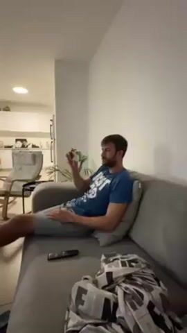 A man lounges on a gray couch, casually dressed in a blue t-shirt and shorts. He holds a small, dark object in his hand, which he inspects. A remote control rests on the couch next to him. His feet are propped up on a yellow ottoman. The camera pans to show the rest of the living room. A television screen displays a basketball game with the EuroLeague logo prominently shown. The living room has a minimalist aesthetic with neutral colors, various potted plants, and a coffee table. A glimpse of a kitchen is visible in the background, suggesting an open-plan layout. Several suitcases and a dining area sit behind the living room.