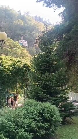 The video shows a sunny outdoor scene with lush green trees and bushes. A cable car is visible ascending a hillside in the background. In the foreground, a lamppost with three globes stands next to a dense bush. There are people walking in the middle ground, and a person is sitting on a bench in the far background. The overall impression is that of a park or recreational area.