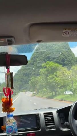 This is a first-person perspective video recorded from inside a vehicle, likely a van or bus, as it drives along a road. The camera is positioned to capture the view through the windshield. In the foreground, hanging from the rearview mirror, is a string of orange flowers and a red ribbon. Below that, a bottle of water is visible, along with part of the vehicle's dashboard, including a screen.

The road ahead curves to the left and is bordered by trees and vegetation. On the right side of the road, there are streetlights. Dominating the background is a massive, lush green mountain covered in trees, its peak obscured by a cloudy blue sky. The sunlight suggests it is daytime. The overall impression is a scenic drive through a tropical or subtropical region.