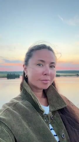 A woman is taking a selfie video outdoors at sunset. She has long, dark brown hair and is wearing a green corduroy jacket over a striped shirt. The background shows a body of water and a colorful sky with hues of orange, pink, and blue. She turns her head to the side to look at the sunset.