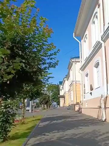 The video shows a street scene with buildings and trees on a sunny day. The camera is moving forward along a sidewalk next to a road. On the right side of the sidewalk, there is a light-colored building with white trim and windows. The building appears to be two stories high. On the left side of the sidewalk, there is a grassy area and a large tree with green leaves. Beyond the grassy area, there is a road with a few cars and a white van. In the background, there are more buildings and trees, and the sky is a clear blue.