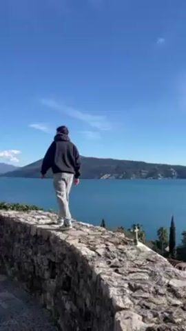 A man in a black hoodie and gray sweatpants stands on a stone wall, overlooking a body of water with mountains in the background. The sky is bright blue with a few clouds. A small stone structure is visible to the left of the man. The man turns towards the camera and gestures towards the landscape. The camera captures the scene from a slightly low angle.