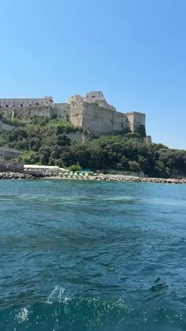 A video captures a view from a boat moving across a body of water. In the background, a large, ancient-looking stone fortress or castle complex sits atop a hill covered with lush green trees. The fortress has multiple levels and sections, with a commanding presence over the landscape. In the foreground of the fortress, along the shoreline, there is a small beach area with umbrellas and what appears to be a restaurant or beach club. Closer to the water's edge, a red lighthouse stands on a rocky breakwater. The water is a deep blue-green and is somewhat choppy, indicating the boat is in motion. The sky is clear and bright blue, suggesting a sunny day. The camera appears to be panning slightly, following the coastline and the fortress as the boat travels.