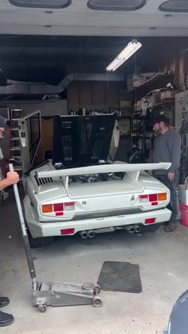 In a garage with a metal garage door, two men stand near the rear of a white Lamborghini Countach. The car's engine cover is open, and the rear spoiler is visible. A silver hydraulic jack sits on the floor near the car. The man on the left is wearing a dark hat, glasses, jeans, and black shoes and operates the jack. The man on the right wears a dark baseball cap, a gray long-sleeved shirt, jeans, and work boots. Shelving lines the back wall, holding various containers and tools. A ladder leans against the left wall, and a red bucket is on the right.