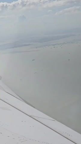 The video shows an aerial view from an airplane window, looking out over a vast expanse of water. The wing of the airplane is visible in the foreground, taking up the bottom left portion of the frame. The water is a muted gray-blue color, and appears somewhat hazy. Scattered across the water are numerous ships, appearing as small, dark specks. In the distance, the land is visible as a faint, light-colored horizon line, with clouds forming overhead. The overall impression is one of distance and scale, with the ships appearing insignificant against the vastness of the sea and sky.