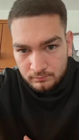 A close-up shot shows a man with a beard and light eyes looking down at the camera. He is wearing a black hoodie. The background is out of focus and appears to be a plain white wall.
