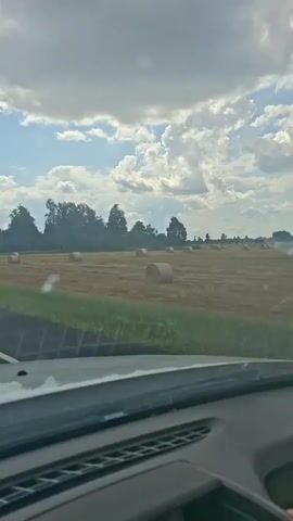 The video shows a view from inside a vehicle, looking out at a rural landscape. The sky is partly cloudy with a blue sky visible. In the foreground, the dashboard of the vehicle is visible. In the middle ground, there is a field with hay bales scattered across it. Trees line the edge of the field in the background. The vehicle appears to be moving on a road as the scenery changes. There are a few traffic signs visible on the side of the road.