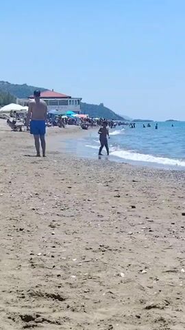 The video shows a sandy beach with people enjoying the water and sun. In the foreground, a man in blue shorts walks away from the camera, his back to the viewer. Further down the beach, a child in dark clothing runs towards the water. The water is blue and appears calm with gentle waves. There are several beach umbrellas and tents set up, and a building with a red roof is visible in the background. Other people are scattered along the shoreline and in the water, some swimming and others relaxing. The sky is clear and blue, indicating a sunny day.