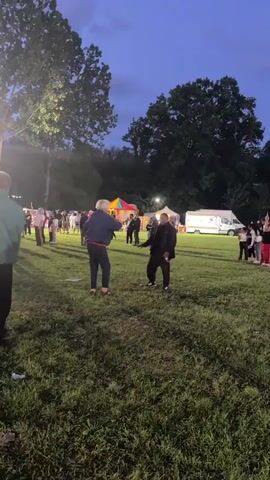 Two people dance on a grass field in an outdoor venue. The person on the left wears a blue jacket, red pants, and slides. The person on the right wears a black jacket and pants with sneakers. Other people, tents, food trucks, and a carousel are in the background. The event takes place at dusk.