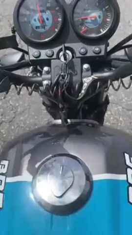 The video shows the view from the front of a motorcycle as it moves down a paved road. The camera is positioned just above the motorcycle's handlebars, capturing the dashboard instruments and the road ahead. The dashboard features two round gauges, one showing the speedometer, and the other presumably the tachometer. The speedometer needle is visible, indicating the speed. A phone mount is attached to the handlebars on the left.  The motorcycle has a two-tone gas tank, featuring a silver and blue stripe. The road ahead is mostly straight with several cars in the distance. Some trees and buildings are visible in the background, and the sky is partly cloudy.  The video appears to be taken on a sunny day.