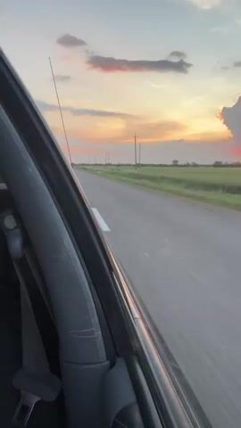 This video shows a view from inside a car driving down a rural road at sunset. The camera is positioned to capture the driver's side of the interior and the road stretching out ahead. The sky is filled with dramatic clouds in shades of orange and pink as the sun sets on the horizon. Telephone poles line the side of the road, and fields of green stretch into the distance. A single car is visible in the distance on the road. The motion blur indicates the car is in motion.