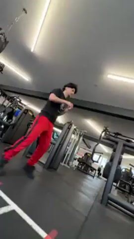 A person wearing a black t-shirt and red pants with a white stripe down the side is standing in a gym. The person is facing to the left and pivoting on their left foot, while throwing punches with their left and right arms in front of them. Various exercise machines are visible in the background, including weight machines and treadmills. The floor is black with a white line painted on it. The lighting is bright, with fluorescent lights on the ceiling.