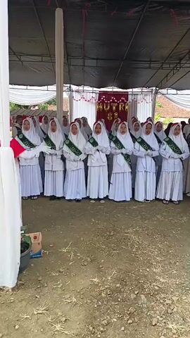 A group of young women in white hijabs and matching white dresses stand in a line, facing forward. They wear green sashes across their chests. A banner with red lettering is visible behind them, and a white tent covers the area. The ground is covered in dirt and gravel.