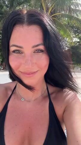 A fair-skinned woman with dark hair, wearing a black bikini and a silver necklace with a swan pendant, is recording a selfie video on a sunny beach. She is smiling at the camera, and occasionally touching her hair, which is blowing in the wind. The background shows a beach with palm trees and other people.