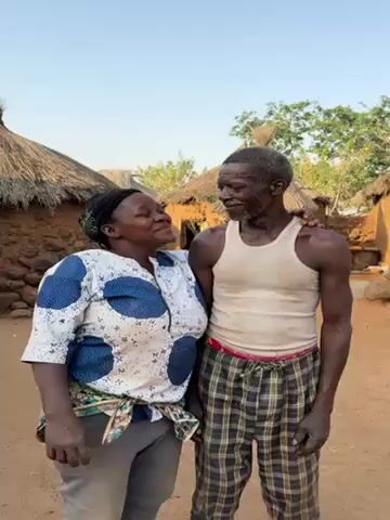 A middle-aged African woman stands close to an elderly African man, smiling up at him. They are both in an outdoor setting, possibly a village, with simple mud brick buildings with thatched roofs visible in the background. The woman is wearing a blue and white patterned shirt, dark pants, and a head covering. The man is wearing a white tank top and plaid pajama pants. The woman has her arm around the man's shoulder. The area around them appears to be a sandy or dirt surface.