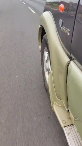 A close-up, side-view video shows the exterior of a dark-colored SUV, possibly a Mahindra Scorpio, as it travels on an asphalt road. The camera is positioned low, near the rear wheel well, capturing the side of the vehicle and the passing road. The SUV has a two-tone paint job, with a lighter beige or tan color on the lower body panels and wheel arches, contrasting with the darker upper body. A small, round orange reflector or light is visible on the rear fender. The vehicle's branding, "Scorpio" and possibly "Pajero" or a similar model name, is partially visible on the side. The road appears to be a highway with a white lane marker. The motion of the vehicle creates a blur effect on the road surface and the surrounding environment, indicating movement. The weather appears to be overcast or the lighting is subdued.