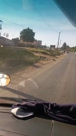 The video shows a view from inside a vehicle driving on a paved road. A person walks on the side of the road, headed in the same direction as the vehicle. Buildings and signs can be seen in the background. The sky is blue. Power lines are visible above. A rearview mirror is visible on the left side of the frame. Part of the vehicle’s dashboard is visible at the bottom of the frame.
