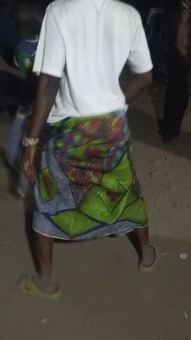 A person dances on a dirt surface surrounded by other people. The dancer wears a white t-shirt, a colorful wrap skirt, and sandals. They also wear bracelets on one wrist and a watch on the other. The setting appears to be a nighttime outdoor event.