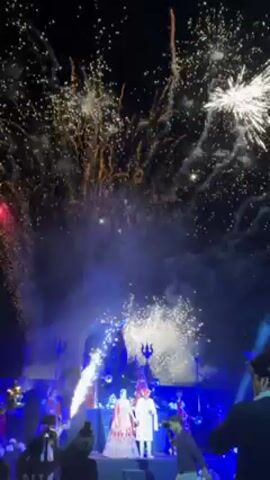 At a night wedding, a couple in traditional Indian wedding garb stand on a stage as fireworks burst in the background. Laser beams and sparklers add to the spectacle. The stage is decorated with flowers and what appears to be Hindu tridents. People stand near the stage, many holding up phones to record the event. The sky is dark, lit up only by the fireworks.