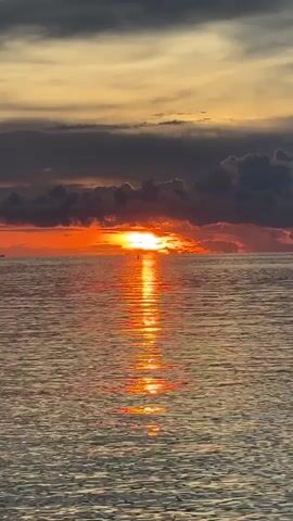 The video captures a vibrant sunset over the ocean. The sun, a bright orange, is partially obscured by a layer of dark clouds along the horizon. The sun's reflection creates a shimmering path of light across the water's surface. The water is calm with small ripples, and the overall sky is a mix of yellows, oranges, and grays. A small, indistinct object can be seen on the horizon near the sun.