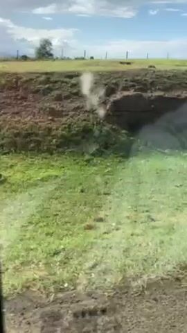 The video depicts a landscape of green grass and dirt hills under a partly cloudy sky, viewed from a moving vehicle through a slightly dirty window. Beyond the grassy area, there's a field with a fence and some buildings in the distance. The camera appears to be in motion, with the scenery passing by quickly.