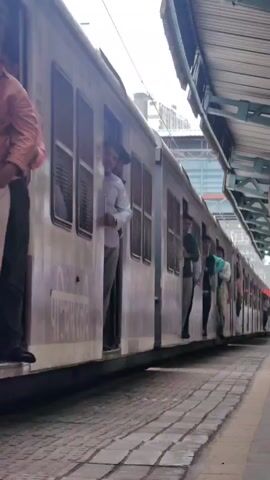 The video shows a train stopped at a station platform. People are leaning out of the open train doors. One person in a white shirt and grey pants is looking out of a doorway. Several other people are visible in other doorways, some leaning out and some standing inside. The train car appears to be metallic with multiple windows. The platform is paved with cobblestone-like material. A covered walkway or structure of the station is visible above the train. The overall scene suggests a busy public transport environment.