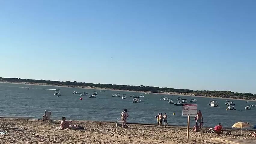 A wide shot captures a sunny beach with people relaxing on the sand and a calm body of water filled with numerous boats in the background. On the sandy beach in the foreground, several individuals are visible. To the left, one person lies on a beach chair, seemingly sunbathing. Further to the right, a group of people stands near the water's edge, including a person in a white top and shorts, and a couple of other individuals in swimwear. A red and white sign is planted in the sand near this group, with text and graphics on it. To the far right, another person sits under a blue umbrella, and further down the beach, other figures can be seen lounging on the sand, some under umbrellas. The water is a deep blue, dotted with many white boats of various sizes, anchored or moving slowly. A distant, tree-covered shoreline stretches across the horizon, with a faint lighthouse visible on the left side. The sky above is clear and a light blue. The overall scene depicts a leisurely day at a popular waterfront location.