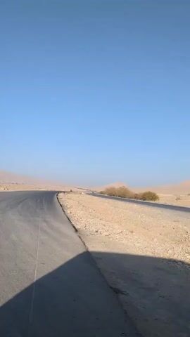 The video shows a desolate desert landscape under a clear blue sky. A paved road stretches into the distance, flanked by rocky, sandy terrain. Sparse vegetation, including small shrubs and a lone tree, dot the landscape. A mountain range is visible in the background. The video seems to be taken from a moving vehicle, casting a shadow on the foreground. There are no people visible.