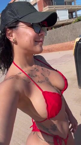 A woman wearing a red bikini and a black baseball cap is taking a selfie on a beach. She is wearing sunglasses and has tattoos on her chest and stomach. The background shows sand, a building, and a trash can. She poses for the camera, puckering her lips.