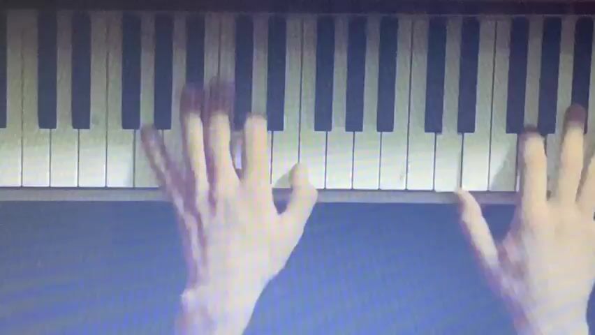 The video displays a close-up, overhead view of a person's hands playing a piano. The keyboard, with its black and white keys, is visible across the frame. The hands are blurred due to motion, indicating active playing. The lighting is somewhat dim, creating a moody atmosphere. The background behind the piano is a dark, solid color. The video captures the movement of the fingers on the keys as the music is played.