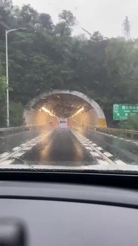 The video shows a first-person perspective from inside a car driving through a tunnel on a rainy day. The car's windshield wipers are active, clearing water and streaks from the glass. The tunnel is illuminated by overhead lights, and the wet road surface reflects the lights and the taillights of vehicles ahead. The tunnel walls are visible, with some signage and markings. The video captures the movement of the car as it progresses through the tunnel, with other vehicles visible in the distance. The overall atmosphere is dark and somewhat hazy due to the rain and the tunnel environment.
