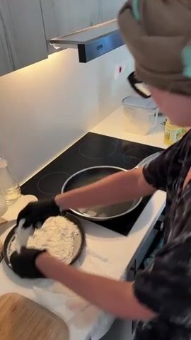 A person wearing black gloves and a bandana is standing in a kitchen. They are in the process of cooking. In front of them is a frying pan with oil in it, and a plate with flour. The person is taking pieces of fish from the plate and dipping them into the flour before placing them into the frying pan. The kitchen appears to be modern with white countertops and a black induction stovetop.