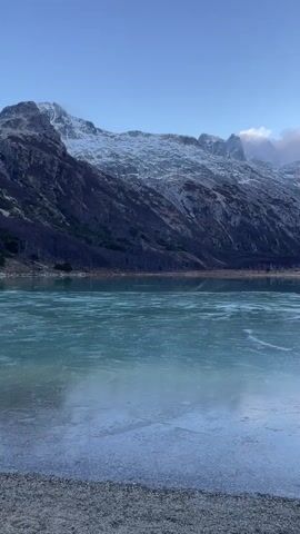 The video shows a serene, partially frozen lake surrounded by mountains, with a rocky shoreline in the foreground. The lake's icy surface has a blue-green hue with visible cracks and patterns. The mountains in the background are a mix of rocky, snow-capped peaks and dark, forested slopes. Some clouds are partially obscuring the mountain tops, and the sky is a clear blue. There are no people visible in the shot.