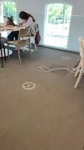The video shows a young woman sitting at a round white table indoors, writing in a notebook. She is wearing a light blue button-down shirt and gray pants. A laptop is on the table in front of her. Other items on the table include a water bottle, phone, pens, and papers. The room has a gray carpet and large, arched windows. Another person is seated at the table, partially visible on the left side of the frame. A white cable lies on the floor.
