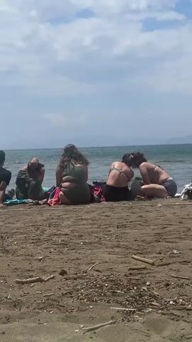 The video shows a group of five people sitting on a beach. The people are all facing the ocean. There is a man in a black shirt sitting on the left side of the frame. Next to him is a child in green shorts who appears to have his head in his hands. To their right is a woman in a green bathing suit. Next to her are two other women in bathing suits. One of the women is facing the other, seemingly engaged in conversation. The beach is sandy and there are some pieces of driftwood scattered around. The ocean is calm and there are islands in the distance. The sky is partly cloudy.