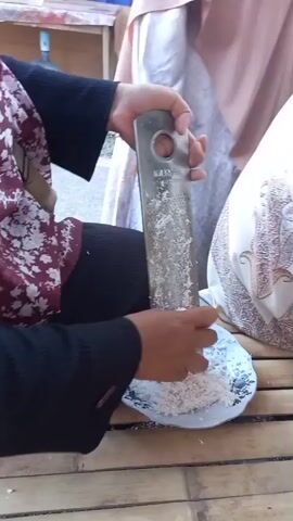 A person is grating coconut with a grater. The person is wearing a maroon and white floral patterned hijab and a black long-sleeved shirt. They are holding a metal grater with a hole in the top and grating a white substance, likely coconut, onto a plate with a blue and white floral pattern. The plate is on a bamboo surface. Another person wearing a light pink hijab is partially visible in the background. The scene appears to be outdoors.