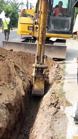 An excavator is digging a trench on the side of a road. The excavator is yellow and operated by a person inside the cab. There are two other people visible, one in a safety vest standing near the excavator, and another person standing on the right side of the frame. The environment appears to be outdoors with some vegetation and buildings in the background. The trench is filled with dirt and rocks.