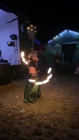 At night, a performer with a flower crown is dancing outdoors with flaming props. The performer is wearing a dark top and a long, flowing green skirt. They are holding two fire props, one in each hand, and spinning them in circles, creating a halo of flames around their head. In the background, there are lights strung across a building, and a few people are seated, watching the performance. The ground is covered in dry grass. There are also speakers and lighting equipment set up to the left of the performer.