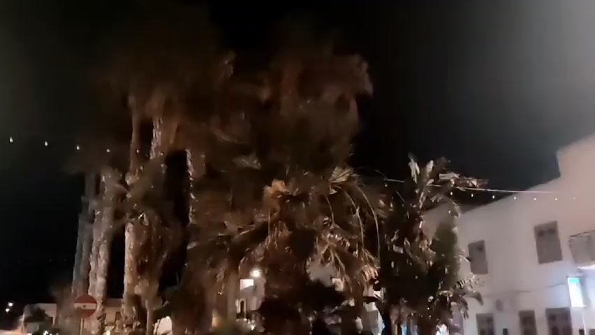 This media file shows a nighttime scene in San Vito Lo Capo, Italy. The footage begins with a view of palm trees and then pans to reveal a street lined with white buildings. The street is illuminated by streetlights and lights from the buildings, creating a warm glow. Several cars are parked along the street, and some people are visible walking in the distance.

The buildings have a Mediterranean architectural style, with arched doorways and balconies. Some of the buildings appear to be shops or restaurants, as suggested by the illuminated signs and open doorways. There are string lights hanging across some of the buildings, adding to the festive atmosphere.

The time of day is clearly night, indicated by the dark sky and artificial lighting. The weather appears to be clear, with no visible rain or fog. The location cues, such as the architecture and palm trees, strongly suggest San Vito Lo Capo, Italy, a coastal town known for its beaches and charming atmosphere.

The overall mood of the video is peaceful and inviting. The gentle illumination and the presence of people suggest a calm evening in a pleasant town. No specific events or activities are prominently featured, but the scene captures the ambiance of a European town at night. There is a brief glimpse of text on a blue sign above an entrance, but it is too blurred to be legible.