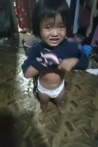 A young child, likely a toddler, is the sole subject of this video. They are wearing a diaper and a dark blue t-shirt with a Captain America shield logo on the front. The child appears distressed and is crying. The setting seems to be an indoor, possibly domestic environment, with a woven mat flooring and what looks like clothes on hangers or a rack in the background. A basket is also visible. The lighting suggests it could be daytime, but the overall environment appears somewhat cluttered. The child is actively engaging with the camera, pulling their shirt up and then down, and looking directly at the viewer with tearful eyes. The child's emotional state is clearly the focal point of the video.