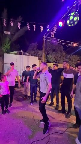 This video captures a lively outdoor gathering at night in Nuseirat, Palestinian Territories. The central subject is a man singing into a microphone, passionately engaged in his performance. He's dressed in a light-colored polo shirt and dark pants, with a distinctive haircut. He appears to be the focal point of the event, drawing the attention of the other people present.

Surrounding the singer are several other men, some of whom are standing close and interacting with him, possibly as friends or fellow performers. One man behind the singer has his arm around him. Others are watching the performance, some with smiles, suggesting a celebratory or festive atmosphere. A young boy in the foreground is also visible, looking towards the singer.

The setting appears to be a somewhat informal outdoor space, likely a courtyard or open area. There are some makeshift structures or walls in the background, along with trees and foliage. Decorative elements, such as strings of lights and paper lanterns, are strung across the area, indicating a festive occasion. A disco ball is visible hanging from a structure, casting colorful lights onto the scene. The ground is covered in gravel or dirt.

The time of day is clearly night, with the artificial lighting playing a significant role in illuminating the scene. The mood seems energetic and joyful, with the singing and the presence of a crowd indicating some form of celebration, perhaps a wedding, party, or community event. The presence of multiple individuals, the music, and the decorations all contribute to a feeling of communal enjoyment. There is no visible text in the video.