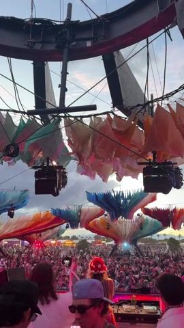 The video shows a crowd of people at a music festival. The scene is brightly lit with pink and red lights, and there are large colorful decorations that look like fabric petals hanging overhead. The perspective is from a stage, looking out at the crowd. There is a DJ performing, and people are dancing and recording the performance. There is some overhead rigging with lights and other equipment. The sky is cloudy in the background.